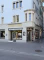 A three-story building with a shop on the ground floor, featuring a sign that reads 'RAHME-LADE'. The shop has large glass windows displaying various items. The building has a modern architectural design with white walls and green window shutters. There a