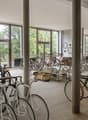 large room with many bicycles parked, wooden floor, large windows, metal poles, tables with bags