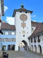 White building with clock tower, multiple windows with flower boxes, archway entrance, cobblestone street, archways along the building