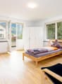 Bedroom: wooden bed, wooden desk with 3 chairs, side table, TV on wall, glass door leading to balcony, white wardrobe, round lamp