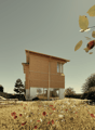 Two-story wooden house with large windows, surrounded by a field of wildflowers and trees. The house appears to be situated in a rural or natural setting with a scenic view.