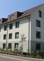 Apartment building with brown roof, many windows, front garden with plants and flowers, a child's playground, and mountains in the background.