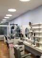 The image shows a room that appears to be a studio or office space. The room has gray walls, a large window, and various shelves and storage units along the walls. There are several mirrors and cosmetic items visible, suggesting it may be used as a beauty