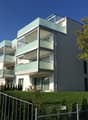 Modern multi-story apartment building with balconies, glass windows, and a landscaped yard with trees and shrubs.