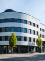 Modern, multi-story office building with a curved facade and glass windows, ArcoWest name sign, trees and cars in front.