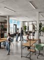 Large open-plan office space with concrete ceiling, floor-to-ceiling windows providing a view, various seating areas including tables, chairs, and lounge furniture, shelving units, and potted plants throughout the space.