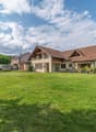 Detached house, brown roof, beige walls, windows with wooden frames, car in the driveway, large green lawn, several bushes and trees, cloudy sky