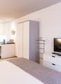Open-plan studio apartment with kitchen, living area, and bedroom. White cabinets and appliances in the kitchen area. Large TV stand with drawers. Patterned rug on the floor.