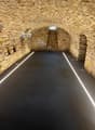A long, dark tunnel with a paved floor and stone walls. The tunnel appears to be underground or within a building, with arched ceilings and lighting fixtures visible.