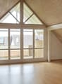 Spacious attic room with vaulted ceiling, wooden paneled walls, large windows providing ample natural light, and access to a balcony or terrace.