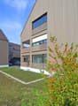 3-story modern building with wooden siding, large windows, and a balcony. The building is surrounded by a grassy area with decorative plants and shrubs.