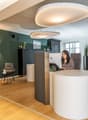 Modern and stylish office space with a reception desk, comfortable seating area, and various storage and display units. The space features hardwood flooring, large windows, and contemporary lighting fixtures. A dishwasher is visible, indicating the presen