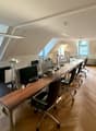 Long desk with multiple computer monitors, office chairs, lamps, wooden floor, white walls, windows, radiators, potted plant