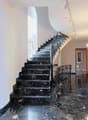 The image shows a grand staircase with black marble steps and an ornate wrought-iron railing. The walls are white, and there is a stained glass window with colorful geometric patterns. The hallway has a polished marble floor and wooden doors leading to ot
