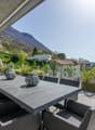Spacious outdoor patio with a large concrete dining table and chairs, surrounded by potted plants and greenery. The patio overlooks a scenic mountain landscape in the background.