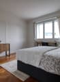 Bedroom with wooden floor, white walls, bed, wooden side table, desk, chair, window with curtains, rug, and potted plants.