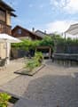 large gravel area, two white umbrellas, a few chairs, a table, plants, trees, trampoline, garden furniture