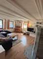 Wooden floor, beige walls, large windows, fireplace, living room couch, coffee table, TV, side table, rose in vase, desk, flower vase, basket on shelves, decorative items on shelves