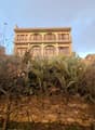 Two-story building with arched windows and balconies, surrounded by a stone wall and cactus plants in the foreground.