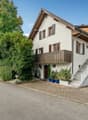 Detached house, 3 floors, white painted walls, brown roof, brick driveway, wooden garage door, brown balcony, plants, shrubs