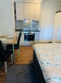 The image shows a studio apartment with an open-plan layout. The kitchen area features white cabinets, a built-in oven, and a dishwasher. There is a dining table with chairs near the kitchen. The living area has a bed with a floral patterned bedding. The