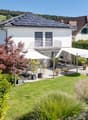 A modern white house with a roof-mounted solar panel system, a spacious backyard with outdoor seating and a jacuzzi, surrounded by greenery.