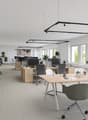 Spacious open-plan office with modern, minimalist design. Large windows provide natural light, and the space is furnished with wooden desks, black office chairs, and hanging light fixtures. Plants and decor elements add a touch of greenery and warmth to t