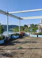 The image shows an outdoor terrace or balcony area with a view of the surrounding landscape. The terrace has a concrete floor and is covered by a metal frame structure. There are several potted plants and flowers placed around the terrace, adding greenery