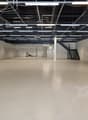 A vacant commercial space with gray concrete flooring, white walls, exposed beams, and overhead fluorescent lighting. There is a staircase in the right corner and an entrance at the back.