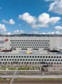 Large modern building with a distinctive white facade featuring a repeating pattern of circular openings. The building appears to be situated in an urban area with a view of the surrounding cityscape. There is a green landscaped area in the foreground of