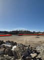 Large open construction site with piles of rocks and debris, construction equipment such as a bulldozer, and temporary orange barriers lining the perimeter. The site is surrounded by buildings and has a clear blue sky overhead.