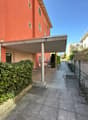 A covered parking area at the front of a building, tiled ground, fenced boundary with greenery, metal railings, multiple windows on building, side entrance