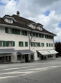 Building with shops on the ground floor, multiple windows with green shutters, chimney, awnings over windows, street sign, parked car
