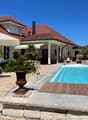 Swimming pool with wooden deck, surrounded by brick paving, patio with lounge chairs, and a large patio umbrella. There are also plants and flowers, and a wooden lounge chair with a canopy on the right side.
