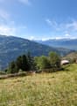 The image shows a scenic mountain landscape with rolling hills, lush green vegetation, and a small village nestled in the valley below. The sky is clear and blue, with wispy clouds. The mountains in the distance appear snow-capped. There is a fence or wir