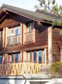 2 story wooden chalet, brown shingles, multiple glass windows, balcony with wooden railings, green trees in front