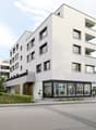 Modern white building, multiple windows, ground floor with glass walls, balcony on the second floor, greenery around