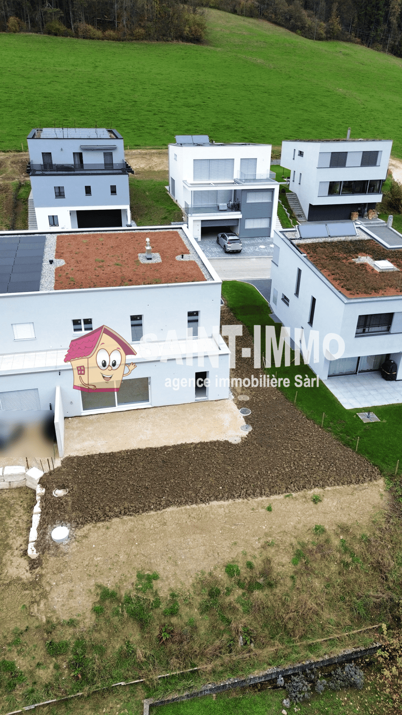 The image shows three modern, white houses with flat roofs and garages. The houses are situated on a grassy field with a forested area in the background. There is a parking space in front of the houses. The image also shows a real estate company logo on o