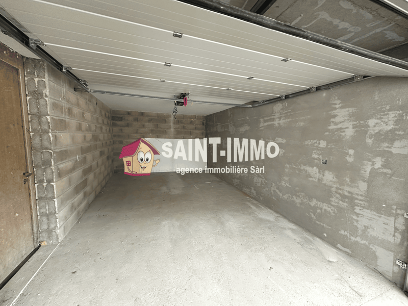 The image shows a garage space with a concrete floor and brick walls. The ceiling is covered with a white paneled system. The Saint-Immo real estate agency logo is prominently displayed on the wall.