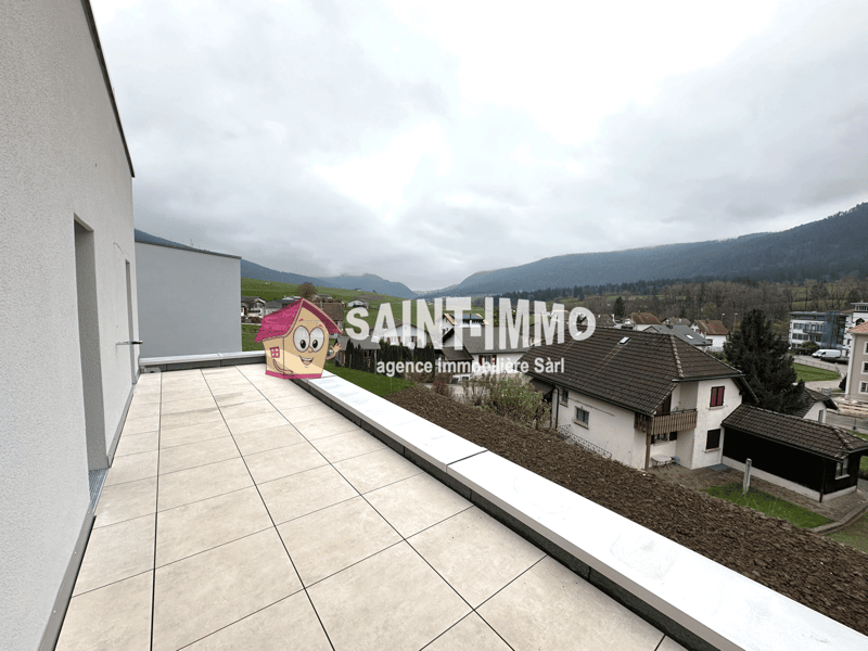 Apartment with a balcony, overlooking a residential area with houses and mountains in the background. The image shows the exterior of the building with the Saint-Immo real estate agency logo visible.
