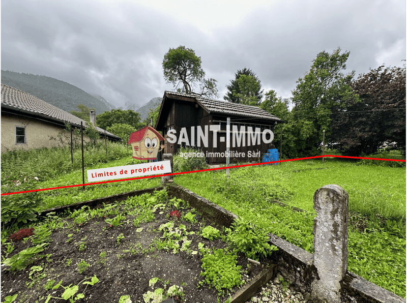 Property surrounded by trees, mountainous landscape in the background, single building with red roof, grassy field, stone post, watermark text