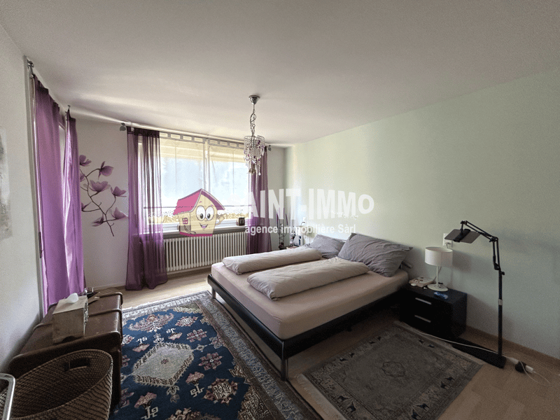 Spacious bedroom with large windows, gray bedding, a crystal chandelier, and a colorful patterned rug. The room also features purple curtains and a decorative wall art piece.