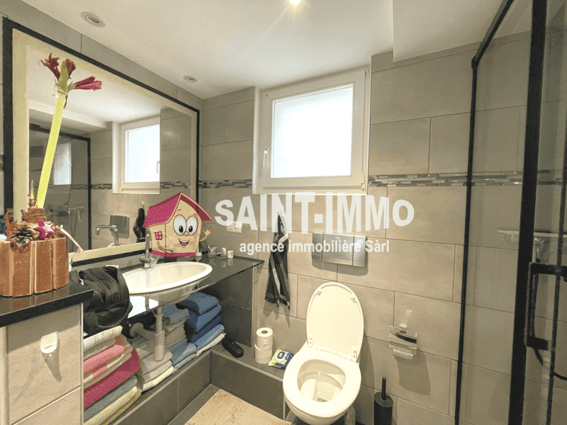 Tiled bathroom with a vanity sink, toilet, and a cartoon house-shaped object on the counter. The walls have the 'Saint-Immo' real estate agency logo printed on them.