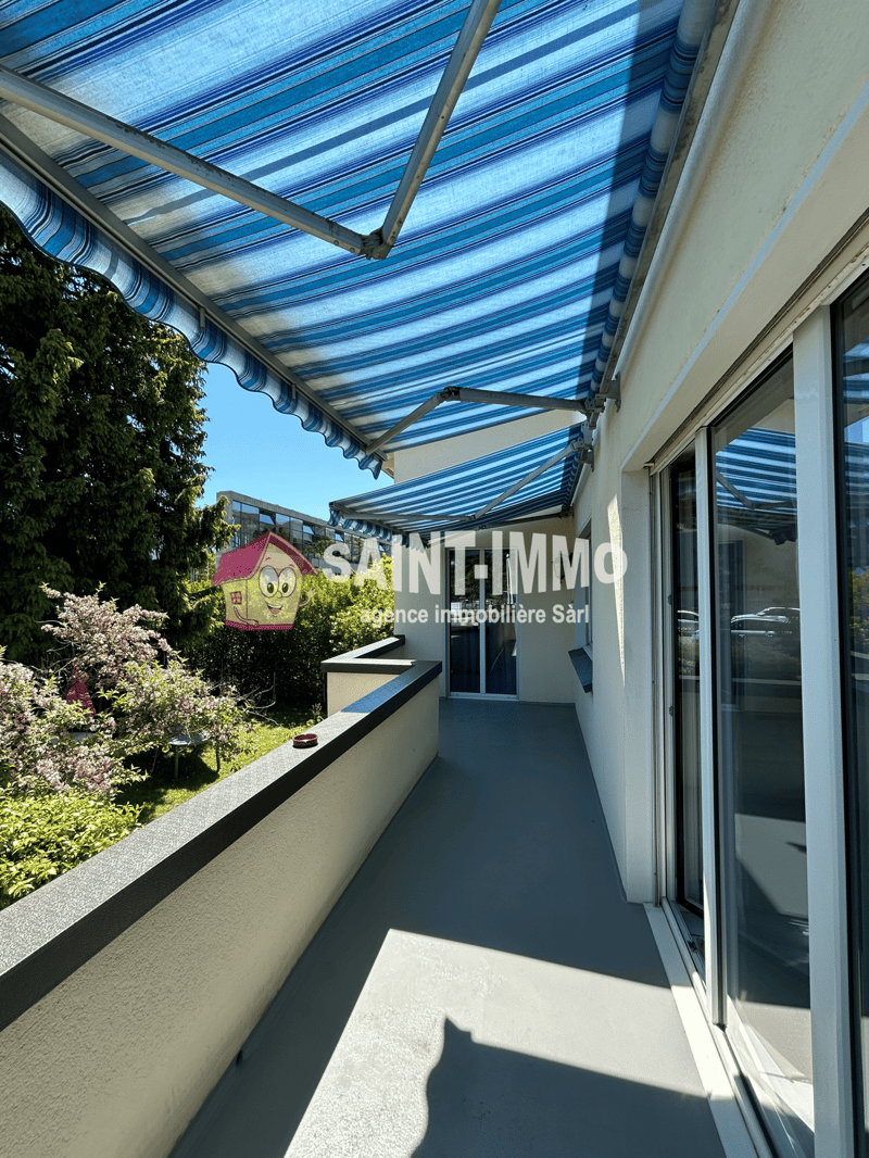 The image shows a balcony or terrace area with a striped blue and white awning covering it. The balcony has a concrete floor and overlooks a garden with trees and shrubs. There are large windows on the adjacent building, and a small decorative house-shape