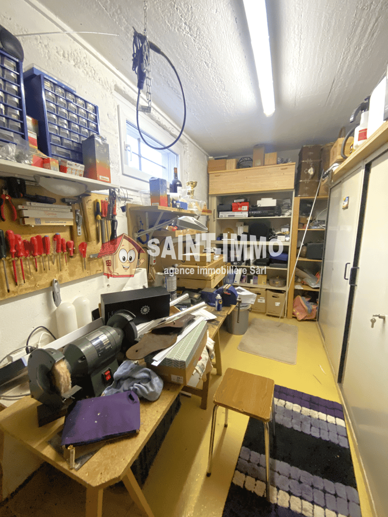 The image shows a cluttered storage room or workshop space. There are various tools, equipment, and boxes stored on shelves and tables. The room appears to be used for storage and organization of various items.