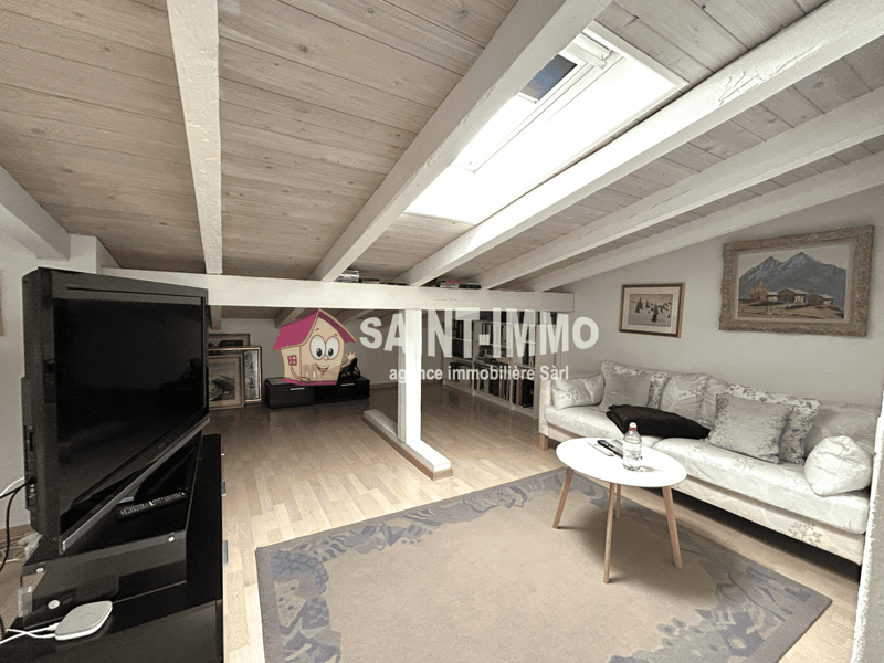 Spacious attic living room with wooden ceiling beams, large windows providing natural light, hardwood flooring, a sofa, coffee table, and a TV stand. The room has a cozy and modern feel with decorative elements like framed artwork on the walls.