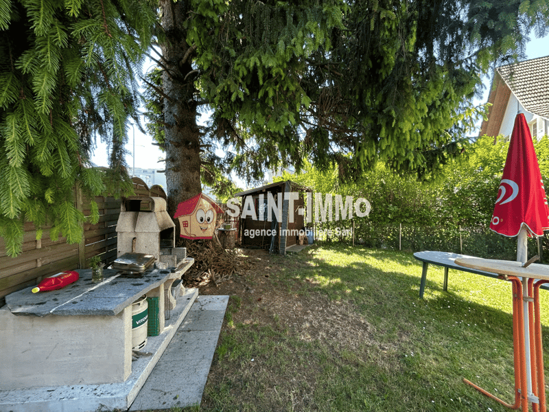 Backyard with outdoor kitchen setup, including a brick oven and grill. There is also a table and chairs, as well as a Saint Immo real estate agency sign. The backyard is surrounded by lush greenery, including tall trees and shrubs.