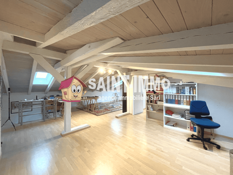 Attic office space with wooden beams, hardwood floors, shelving units, and a large window providing natural light. The space appears to be used as a workspace, with a desk and chair visible.