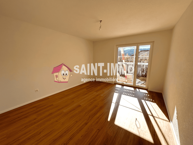 Empty room with hardwood flooring, beige walls, and large windows providing natural light. The room appears to be part of a property listed by the real estate agency 'Saint-Imier'.