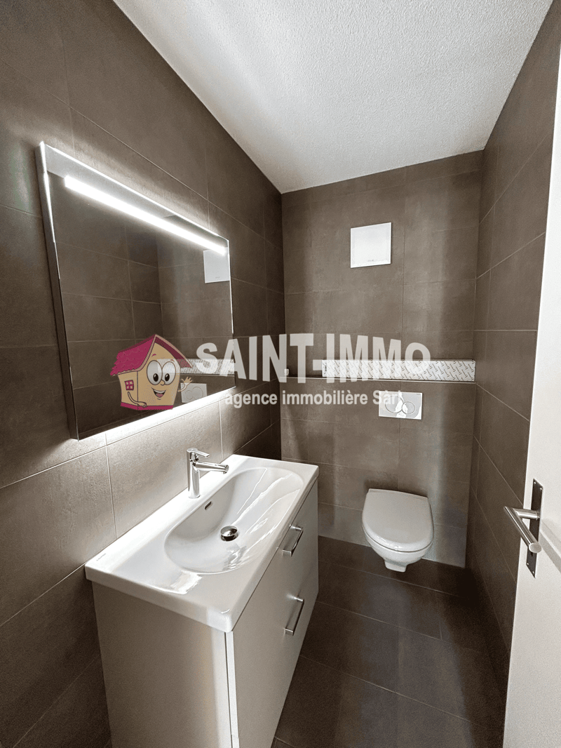 The image shows a modern bathroom with gray tiled walls, a white vanity with a sink, and a toilet. The bathroom appears to be well-lit with a mirror on the wall.